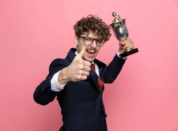 elegantní mladý podnikatel v námořnické modré oblek nosí brýle, takže palce nahoru znamení a drží trofej na růžovém pozadí - Fotografie, Obrázek
