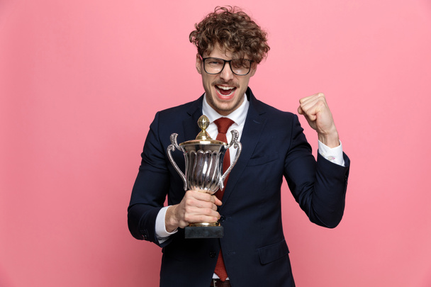 joven emocionado en traje azul marino con gafas y gritando, sosteniendo trofeo, sosteniendo puños y celebrando la victoria sobre fondo rosa - Foto, imagen
