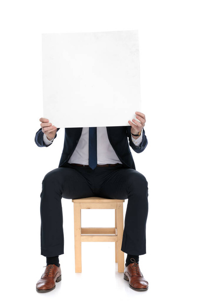 Молодой бизнесмен закрывает лицо пустым рекламным щитом, сидя на стуле на белом фоне студии - Фото, изображение