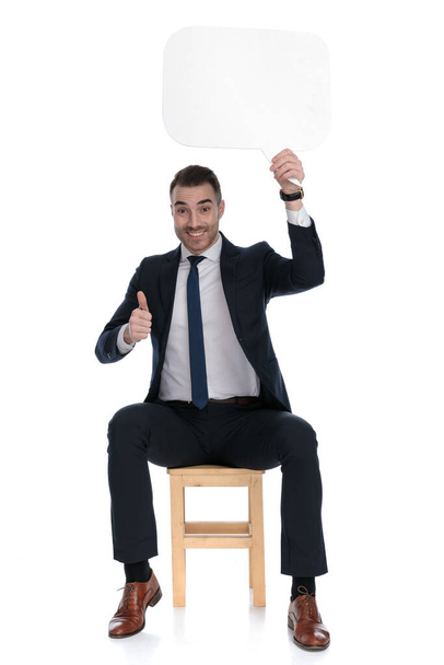 Boldog üzletember tartja beszéd buborék és így hüvelykujj-up, miközben ül egy széken fehér stúdió háttér - Fotó, kép