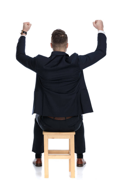 Pohled zezadu na slavícího podnikatele, jak hází ruce do vzduchu a sedí na židli na bílém pozadí studia - Fotografie, Obrázek