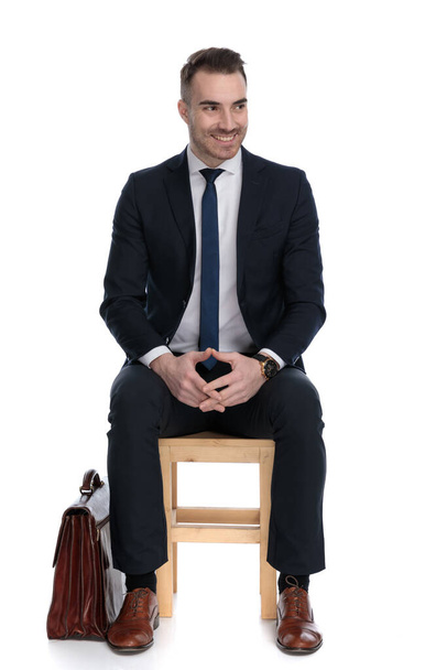 Ενθουσιασμένος επιχειρηματίας παίζει με τα χέρια του και κοιτάζει μακριά δίπλα στο χαρτοφύλακα ενώ κάθεται σε μια καρέκλα σε λευκό φόντο στούντιο - Φωτογραφία, εικόνα