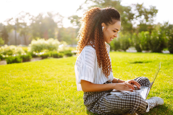 Portrait of pretty young woman sitting on green grass in park with  laptop. Happy woman, student, freelancer with wireless headphones calling on laptop, talk by webcam, video conference. - Foto, Imagen