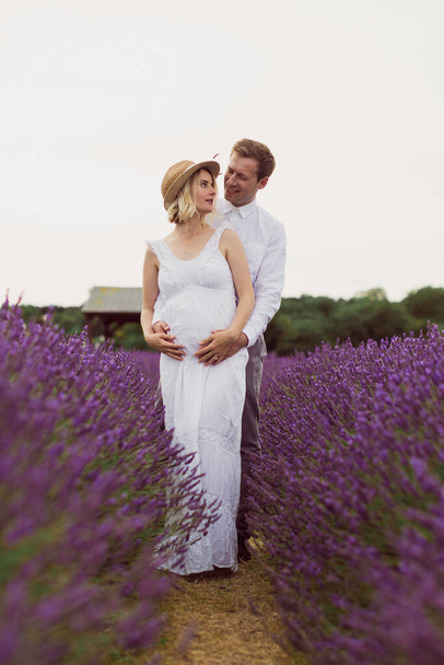 The pregnant wife with her happy husband is standing in a hug in the lavender field and looking at each other - Foto, imagen