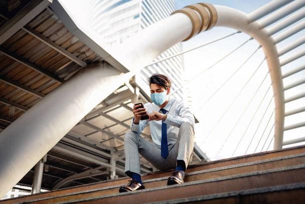Молодой азиатский бизнесмен в хирургической маске и с помощью смартфона в городе. сидя на лестнице. Здравоохранение в новой концепции нормального образа жизни - Фото, изображение