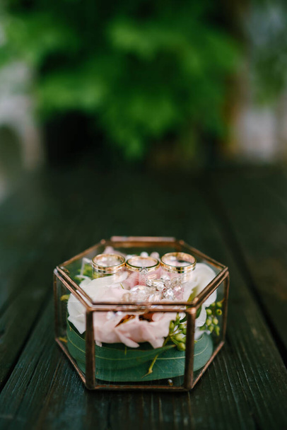 Morsiamen ja sulhasen vihkisormukset ja kihlasormus koristeellisessa lasilaatikossa, jossa on vaaleanpunainen kukka. - Valokuva, kuva
