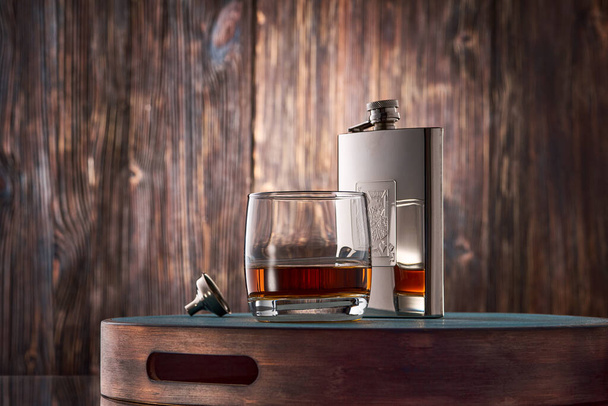 A round glass of whiskey and a metal flask stand on a tray against an old wooden wall. Low key. - Zdjęcie, obraz
