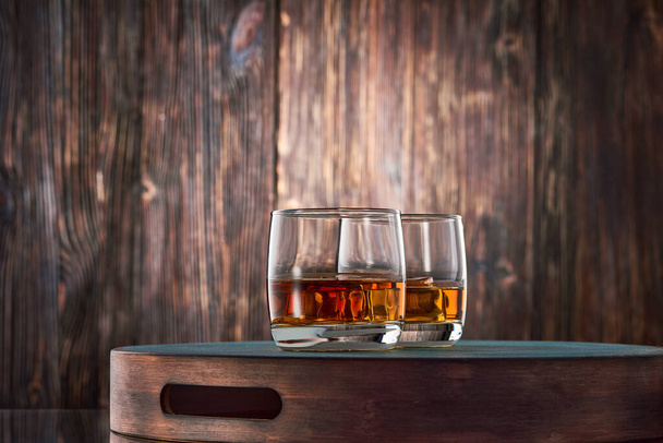 Two round glasses of whiskey on the rocks stand on a tray against an old wooden wall. Low key. - Zdjęcie, obraz