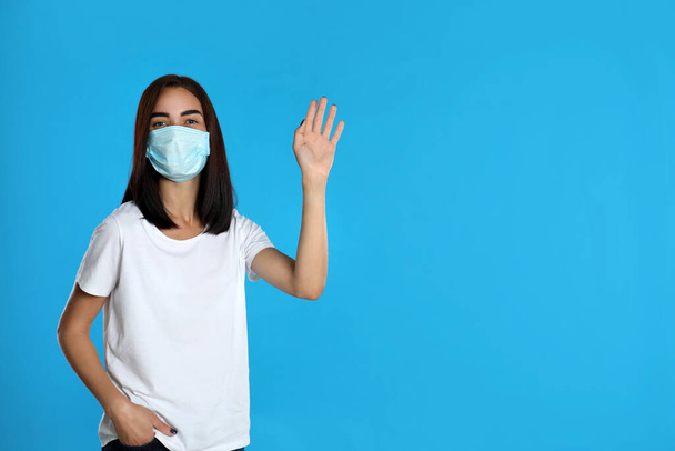 Young woman in protective mask showing hello gesture on light blue background, space for text. Keeping social distance during coronavirus pandemic - Fotó, kép