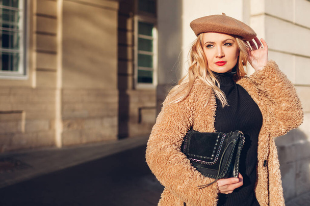 Portrait of stylish young blonde woman wearing teddy coat beret holding purse in city. Autumn fashion accessories. Trendy modern clothes outfit - Photo, Image