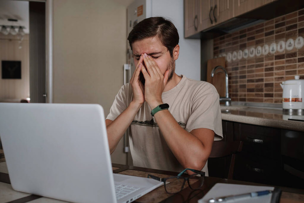 Zmęczony młody człowiek odczuwa ból wzroku trzymając okulary pocierając suche podrażnione oczy zmęczone pracą komputera, zestresowany człowiek cierpi na ból głowy zły wzrok problem siedzieć przy stole w domu za pomocą laptopa - Zdjęcie, obraz