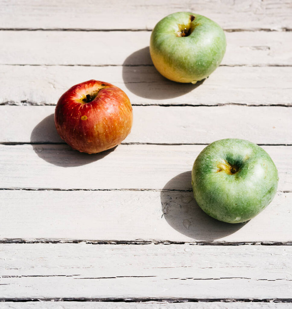 Ένα κόκκινο μήλο και δύο πράσινα μήλα βρίσκονται σε λευκό ξύλινο φόντο. Φρέσκα μήλα στην κουζίνα στο τραπέζι, τρία μήλα. - Φωτογραφία, εικόνα