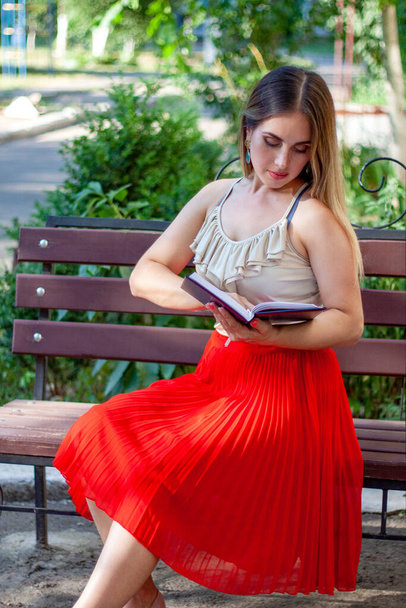 Gyönyörű lány piros szoknyában és fehér felsőben ül a padon és könyvet olvas. - Fotó, kép