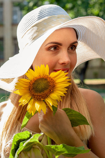 One beautiful girl in a white hat stands in the park with a sunflower in her hand. She looks on the right side and smiles. The weather is warm and sunny - Photo, Image