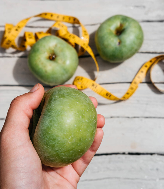 Ihminen koskettaa vihreitä tuoreita omenoita ja keltaista senttinauhaa numeroilla. Herkullinen ja terveellinen ruoka vitamiineilla kuntoa ja terveellistä elämäntapaa varten. - Valokuva, kuva