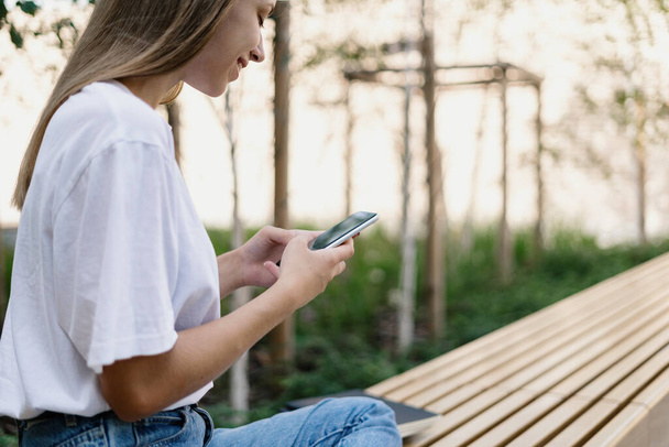 インターネット中毒の概念。現代のスマートフォンでオンラインアプリを使用して笑顔の若い学生の女の子のプロフィール側のビュー,コピースペースとぼやけた背景の近くの木製のベンチに座って - 写真・画像