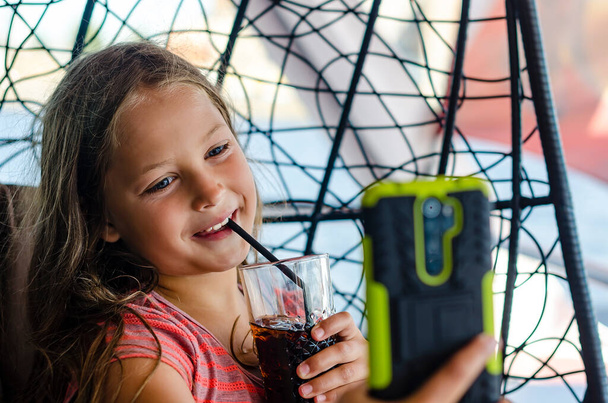 Adorable smiling girl looks at mobile and drinks juice. - Photo, Image