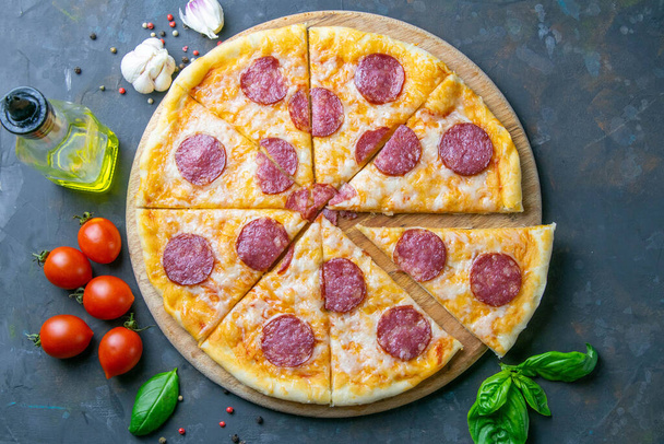 Tasty pepperoni pizza and cooking ingredients tomatoes basil on black concrete background. Top view of hot pepperoni pizza. - Photo, Image