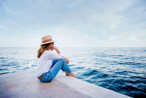 Sahilde oturup deniz manzarasının tadını çıkarırken hasır şapka ve güneş gözlüğü takan bir kadının fotoğrafı.. - Fotoğraf, Görsel