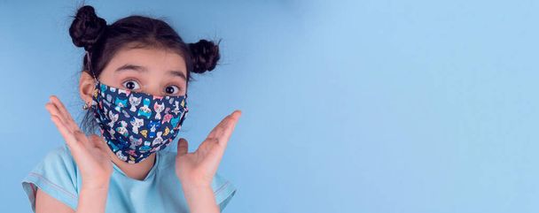 Klein grappig meisje met een kleurrijk masker. Opwinding en fascinatie concept.Coronavirus blauwe achtergrond. dwaze grimmige komische gekke gebaar. Grappige uitdrukking. Covid-19. panorama achtergrond - Foto, afbeelding