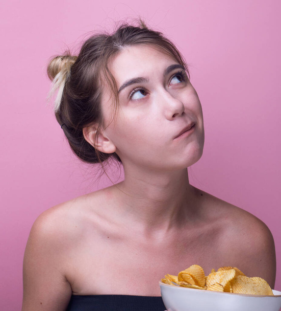 空腹の面白い女性の色のピンクの背景にジャガイモのチップを食べる。魅力的で幸せな女の子揚げジャガイモを食べる。クローズアップ・スタジオ・ポートレート。隔離されてる。不健康なスナック、パノラマ - 写真・画像