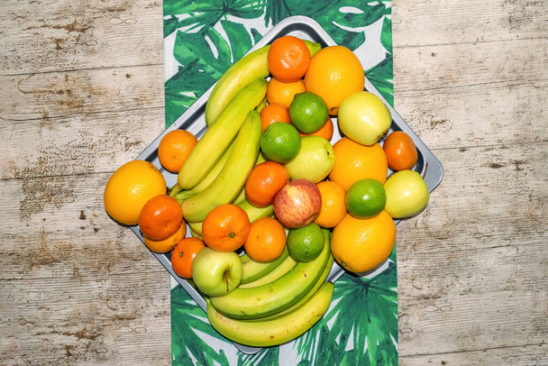 Bandeja antiaderente cheia de bananas lavadas, tangerinas, maçãs e limas com uma perspectiva de cima para baixo em cima de um corredor de mesa de folhas tropicais. - Foto, Imagem