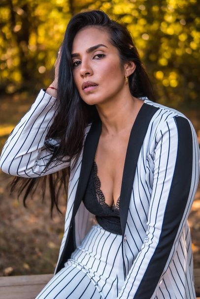 Summer lifestyle, young Latin brunette girl in a very elegant white suit with black stripes. In a beautiful park sitting, portrait stroking her hair - Valokuva, kuva