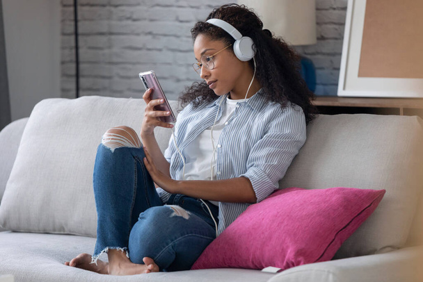 Shot of beautiful young woman listening to music with headphones while using her smartphone sitting on sofa at home. - Foto, immagini