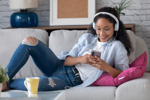 Shot of beautiful young woman listening to music with headphones while using her smartphone sitting on sofa at home. - Foto, imagen