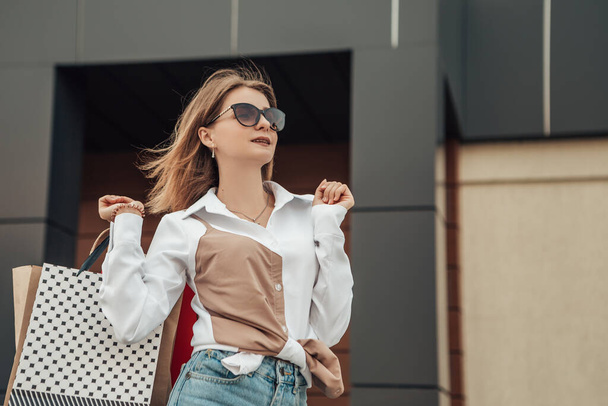 Portret europejskiej pani trzymać za rękę kolorowe torby kobieta po zakupach w okularach słonecznych biały koszula jeansy sklep w pobliżu centrum handlowego na ulicy sprzedaży czarny piątek sezon bokeh tło - Zdjęcie, obraz