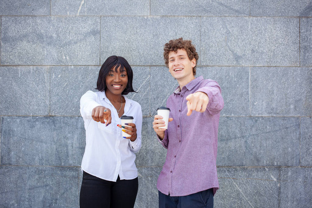 多人種の友人が一緒に、灰色の背景に対して、ポーズを探し、カメラで指を指してハングします。幸せなアフリカの女性と笑顔ヨーロッパの男はコーヒーのカップを保持しています. - 写真・画像