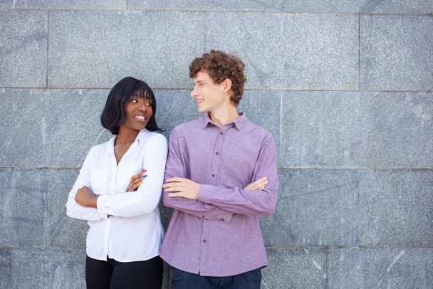 Multiracial male and female friends with crossed arms smiling and looking at each other while standing near wall of building on street - Photo, image
