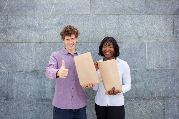 親指を立てたヨーロッパ系の青年と幸せな顔をしたアフリカ系アメリカ人の女性。灰色の背景に外の食品袋を保持する国際的な友人。食と人のコンセプト. - 写真・画像