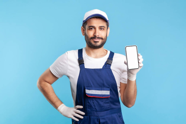 Worker in overalls and gloves holding mobile phone with blank display, device mock up for online delivery order app, house repair maintenance services. indoor studio shot isolated on blue background - Foto, immagini