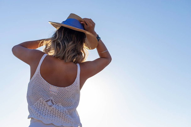 Vista posteriore di una bella donna asiatica che gioca con un sombrero estivo sulla spiaggia al tramonto indossando un elegante abito bianco estivo. Stile di vita donna concetto di libertà viaggiare da soli e rilassanti. - Foto, immagini