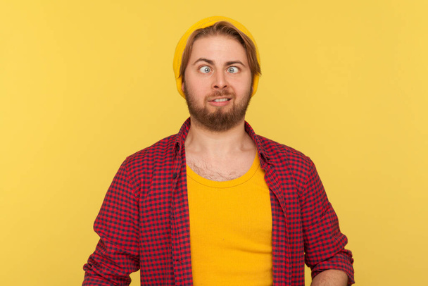 Funny dumb hipster bearded guy in beanie hat and checkered shirt making silly face with eyes crossed, looking at camera stupid brainless expression. indoor studio shot isolated on yellow background - Фото, зображення