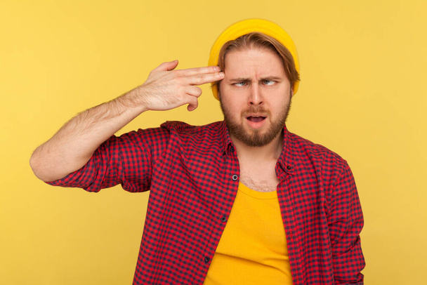 Depressed hipster guy in beanie hat and checkered shirt committing suicide with finger gun gesture, looking cross-eyed funny expression, feeling crazy. indoor studio shot isolated on yellow background - Photo, Image