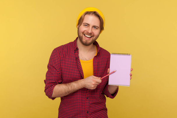 Write reminder to-do list into notebook! Happy hipster trendy guy in beanie hat showing empty paper page, template mock up for creative idea, advertising text. studio shot isolated, yellow background - Φωτογραφία, εικόνα