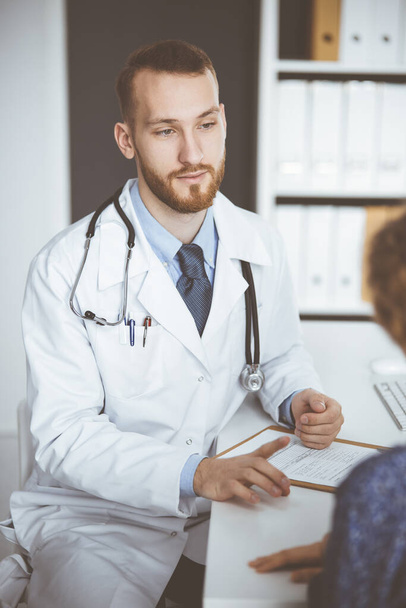 Friendly red-bearded doctor and patient woman discussing current health examination while sitting in sunny clinic. Medicine concept - Photo, image
