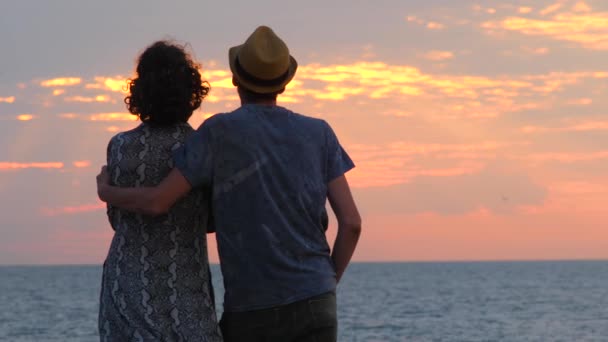 Muž objímá ženu, když se dívá na západ slunce na pláži. Zadní pohled. Manžel v lásce sleduje krásné moře na pobřeží. Romantické pozadí scény. Šťastná rodina se dívá na obzor při západu slunce - Záběry, video