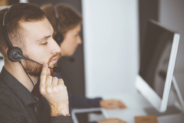 Red-bearded businessman talking by headset near his female colleague while sitting in modern office. Diverse people group in call center. Telemarketing and customer service - Foto, Bild