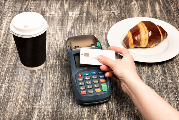Nainen maksaa kortilla. NFC maksupäätteen ja ottaa pois kupin kahvia suklaa croissant puinen pöytä. Luottokortti tai puhelin maksaa pos pankki laite, kortti kone. - Valokuva, kuva