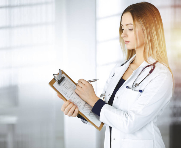 Young optimistic woman-doctor is holding a clipboard in her hands, while standing in a sunny clinic. Portrait of friendly female physician with a stethoscope. Perfect medical service in a hospital - Photo, Image