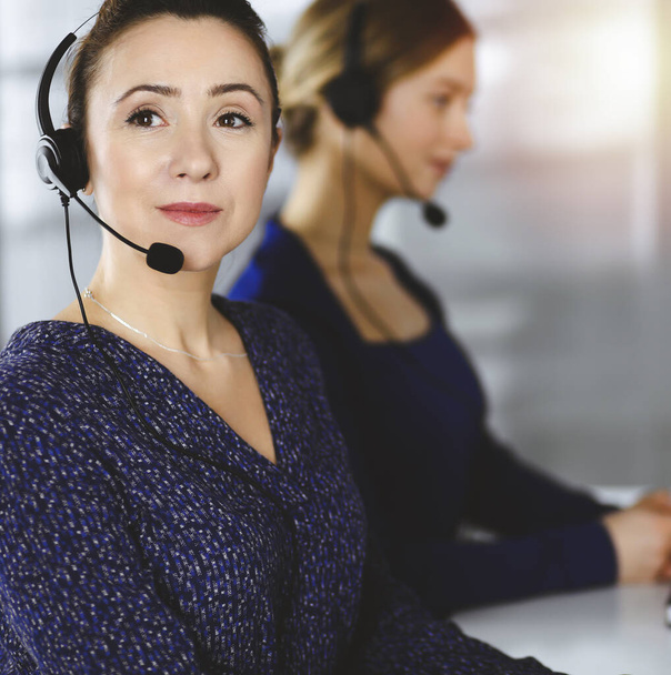 Two busineswomen have conversations with the clients by headsets, while sitting at the desk in a sunny modern office. Diverse people group in a call center. Telemarketing and customer service - Φωτογραφία, εικόνα