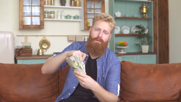 Hombre pelirrojo barbudo comiendo su dinero sentado en el sofá en casa. - Imágenes, Vídeo