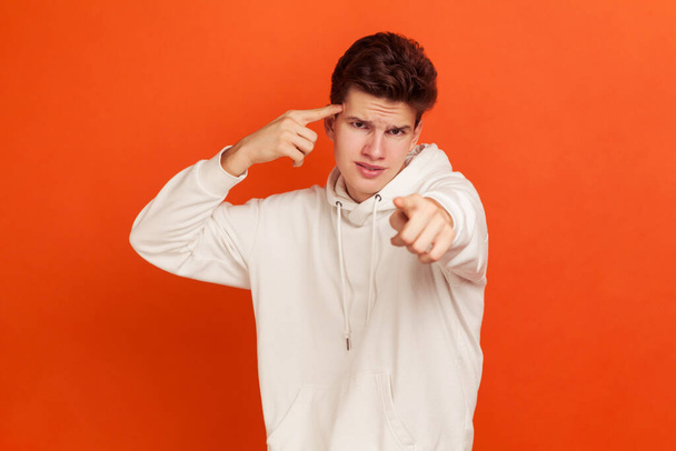 You are stupid! Self confident egoistic teenager holding finger near temple and pointing on you, showing stupid gesture, mocking. Indoor studio shot isolated on orange background - Foto, imagen