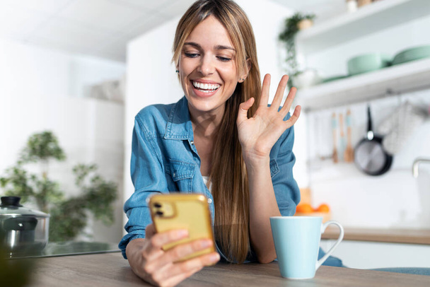 Shot of pretty young woman waving hand while making video call with smartphone during morning tea in the kitchen at home. - Foto, Bild