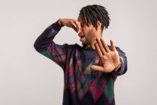 Side view african man with beard and dreadlocks showing no gesture with palm holding breath with fingers on nose, feeling unpleasant smell, disgust. Indoor studio shot isolated on gray background - 写真・画像