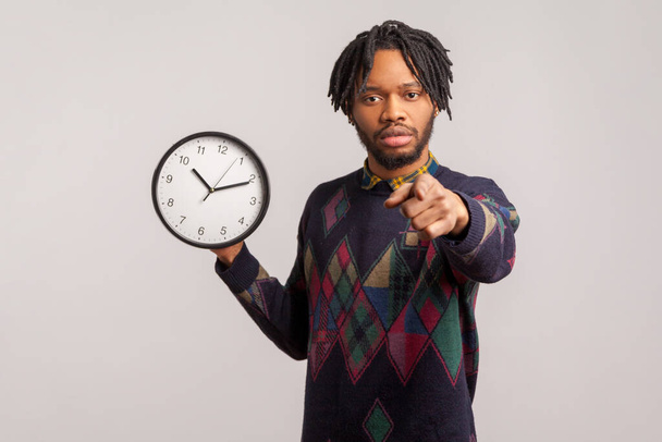 Serious bossy african man with dreadlocks holding wall clock in hand pointing finger at camera with concentrated look, time to action, motivation. Indoor studio shot isolated on gray background - Photo, Image