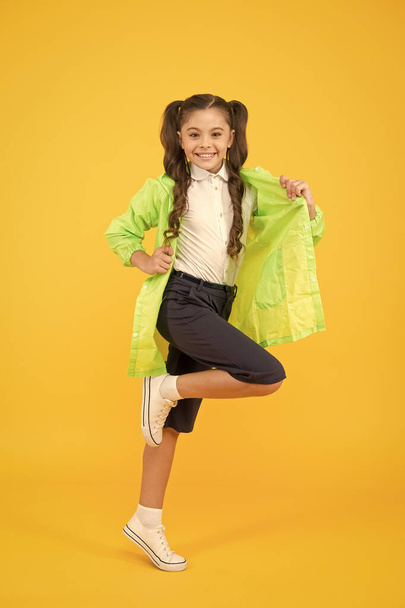 Waterproof concept. Small schoolgirl wear water resistant clothes for rainy day. Cute schoolgirl feel protected for spring weather. Adorable child. Happy schoolgirl wear raincoat on yellow background - Photo, Image